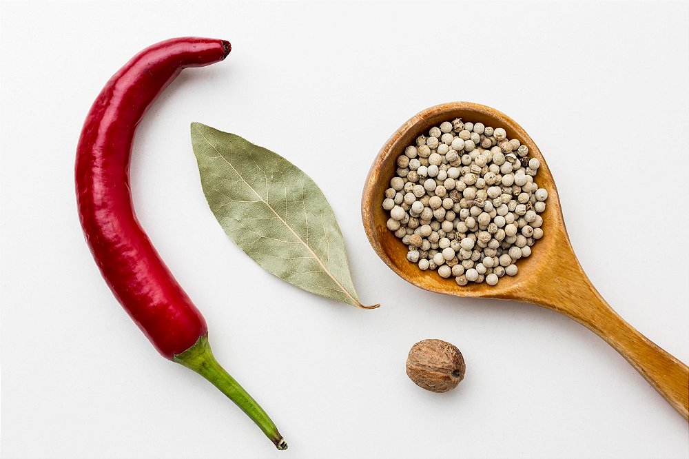 Foto de alimentos termogênicos, como uma pimenta e uma colher cheia de sementes de pimenta, sobre uma mesa branca