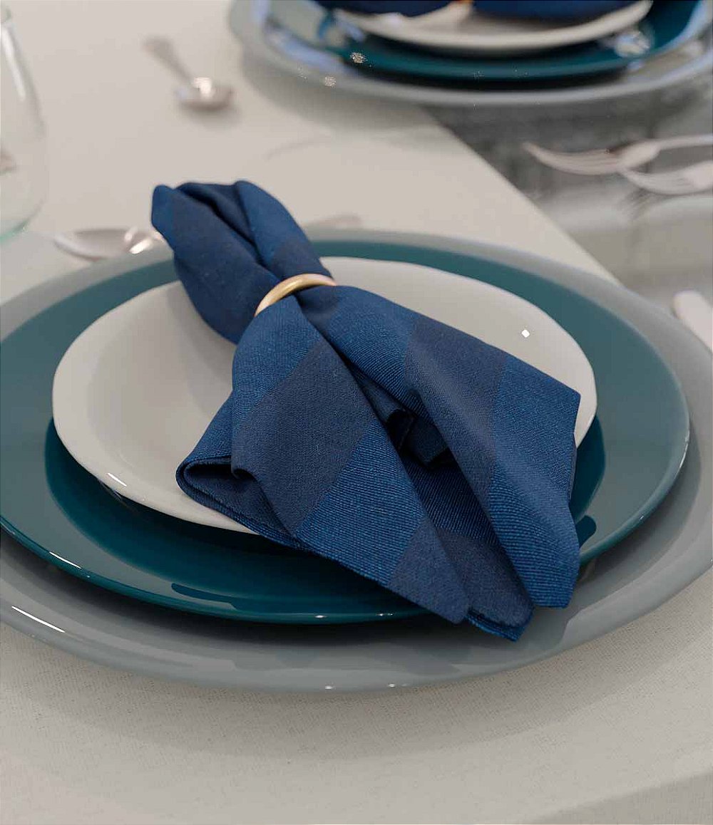 mesa posta com guardanapo azul listrado por cima de prato branco e anel de guardanapo dourado