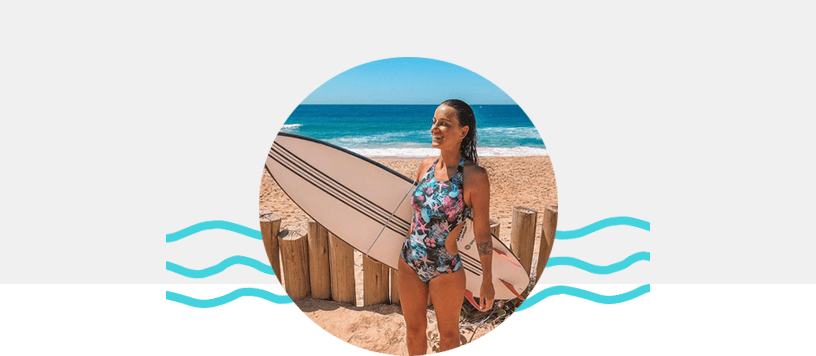 Na imagem a Barbara Speck aparece com os cabelos molhados em uma praia com o mar ao fundo, seguirando uma prancha de surf e vestindo um maio florido da Loveboard