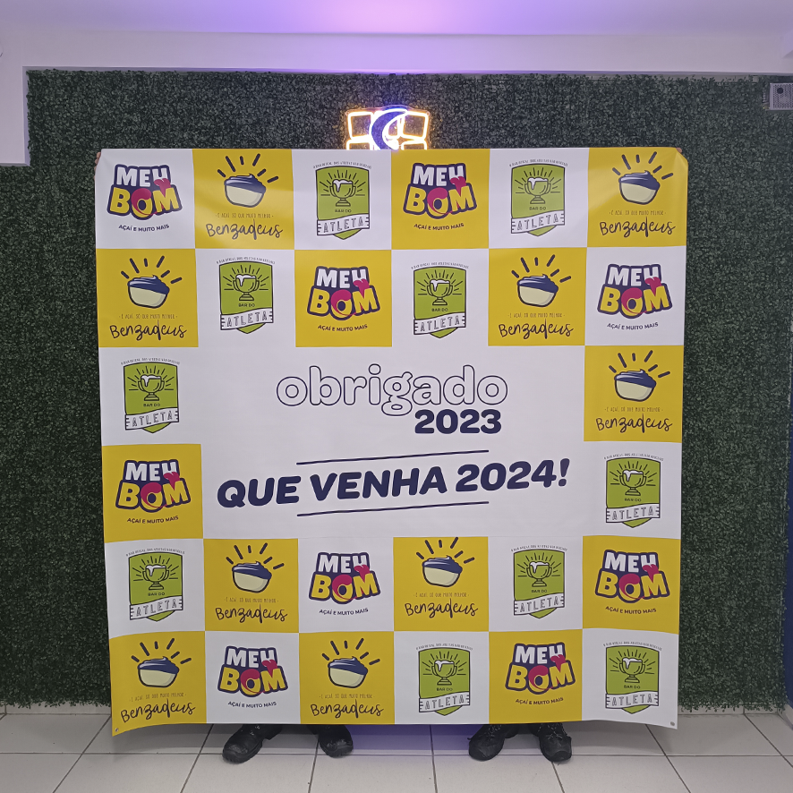 Banner comemorativo dando boas-vindas a 2024, com os logotipos das empresas Benzadeus (amarelo e roxo) e Atleta (verde). O banner está em uma entrada com fundo de plantas artificiais verdes.