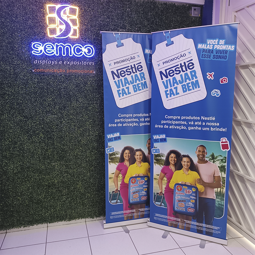 Banner RollUp personalizado da Nestlé (azul) com uma família (pai, mãe e filha) promovendo a campanha 'Viajar faz bem', recomendando ao cliente ir a área de ativação ao comprar produtos Nestlé para receber brindes e concorrer a uma viagem.