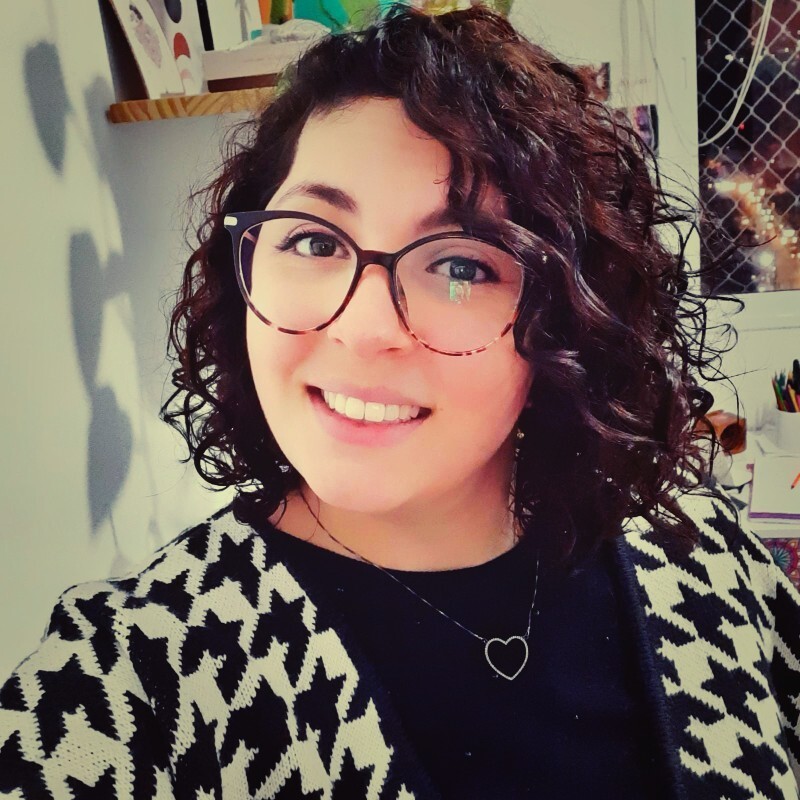 Eu sou uma mulher branca, de cabelo cacheado, castanho e acima dos ombros. Eu uso óculos e estou usando uma blusa preta e branca, com uma base preta e um colar com pingente de coração. Eu estou sorrindo na foto.