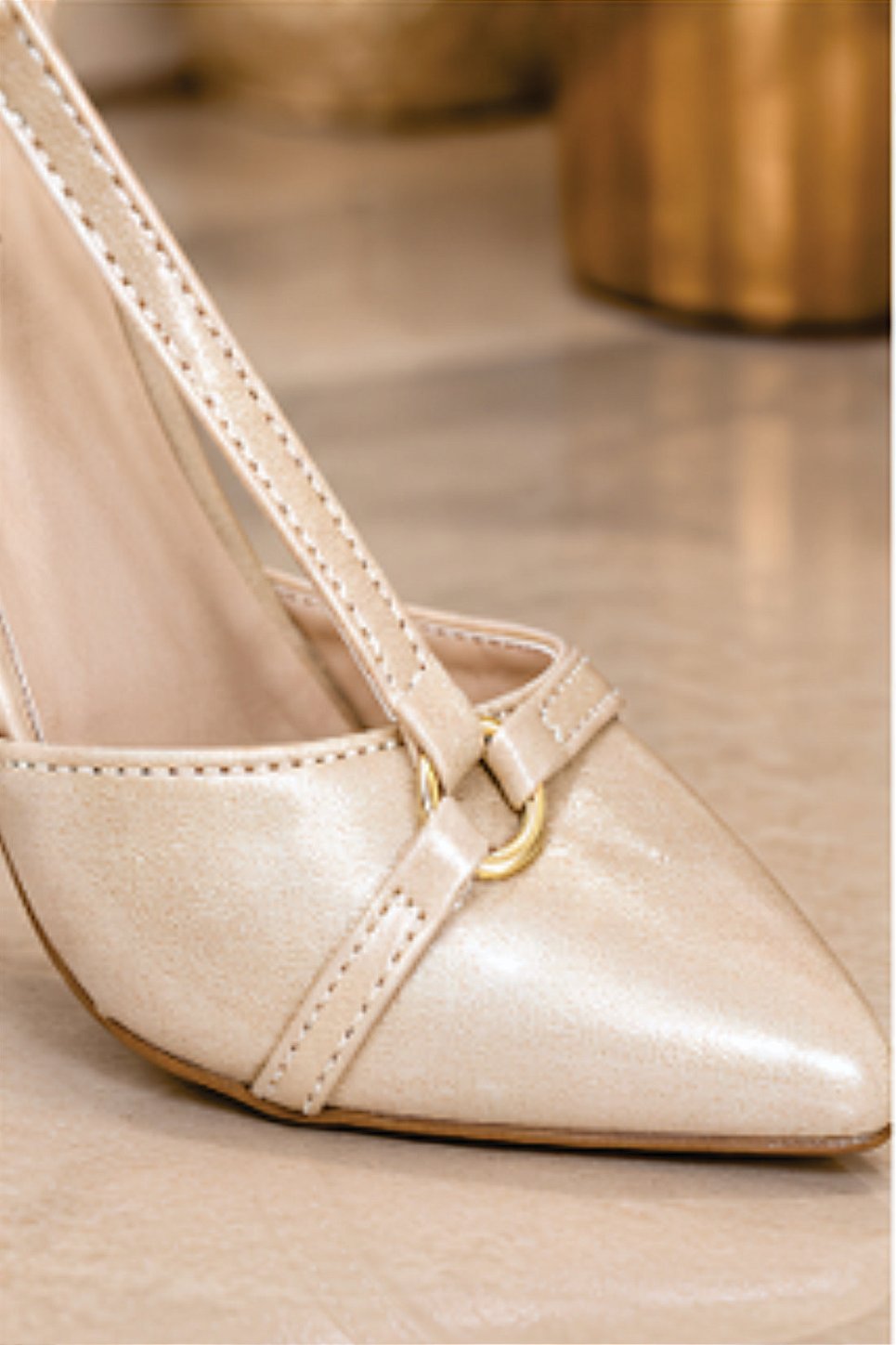Sapato scarpin feminino modelo slingback de bico fino na cor napa Bege Torricella com fivela dourada, exposto sobre uma mesa de vidro em uma galeria de arte moderna luxuosa.