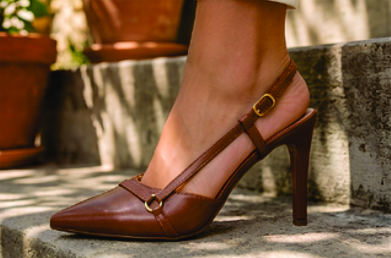 Sapato scarpin feminino modelo slingback de bico fino na cor napa Caramelo Torricella  com fivela dourada, exposto sobre uma mesa de vidro em uma galeria de arte moderna luxuosa.02