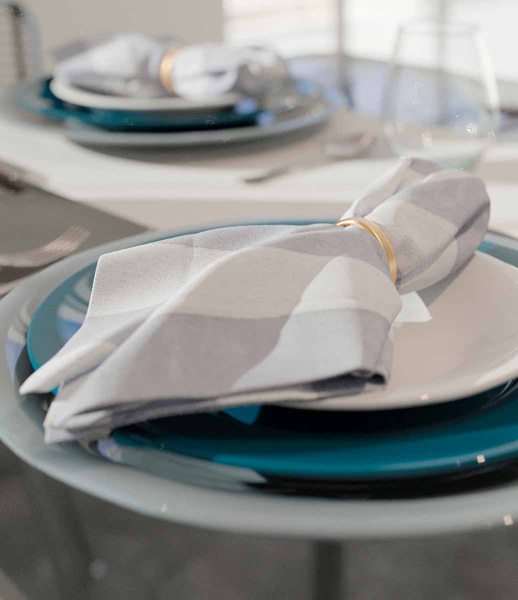 mesa posta com guardanapo cinza listrado dobrado em cima de louça branca e anel de guardanapo