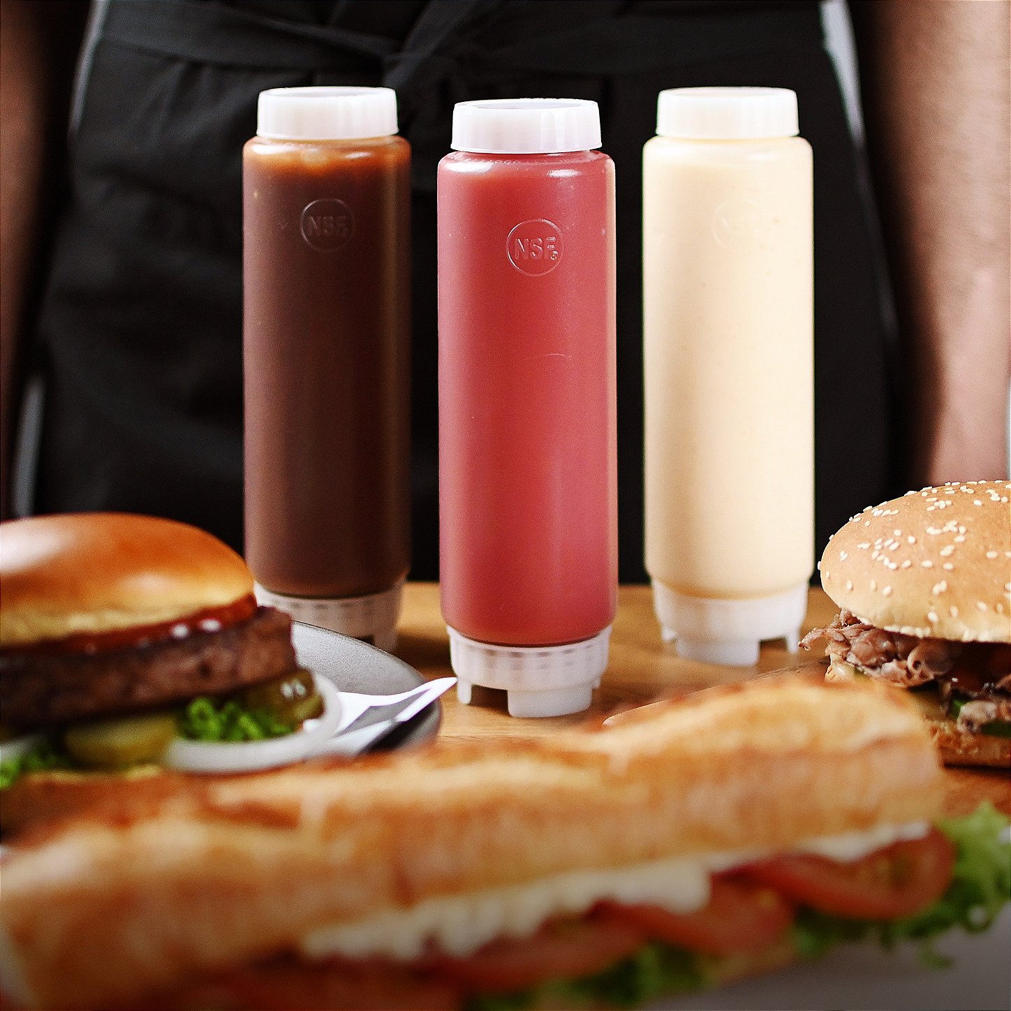 Bisnagas FIFO para molhos com tampas brancas, ideais para ketchup, mostarda e maionese, sobre uma bancada com hambúrgueres e sanduíches ao fundo.