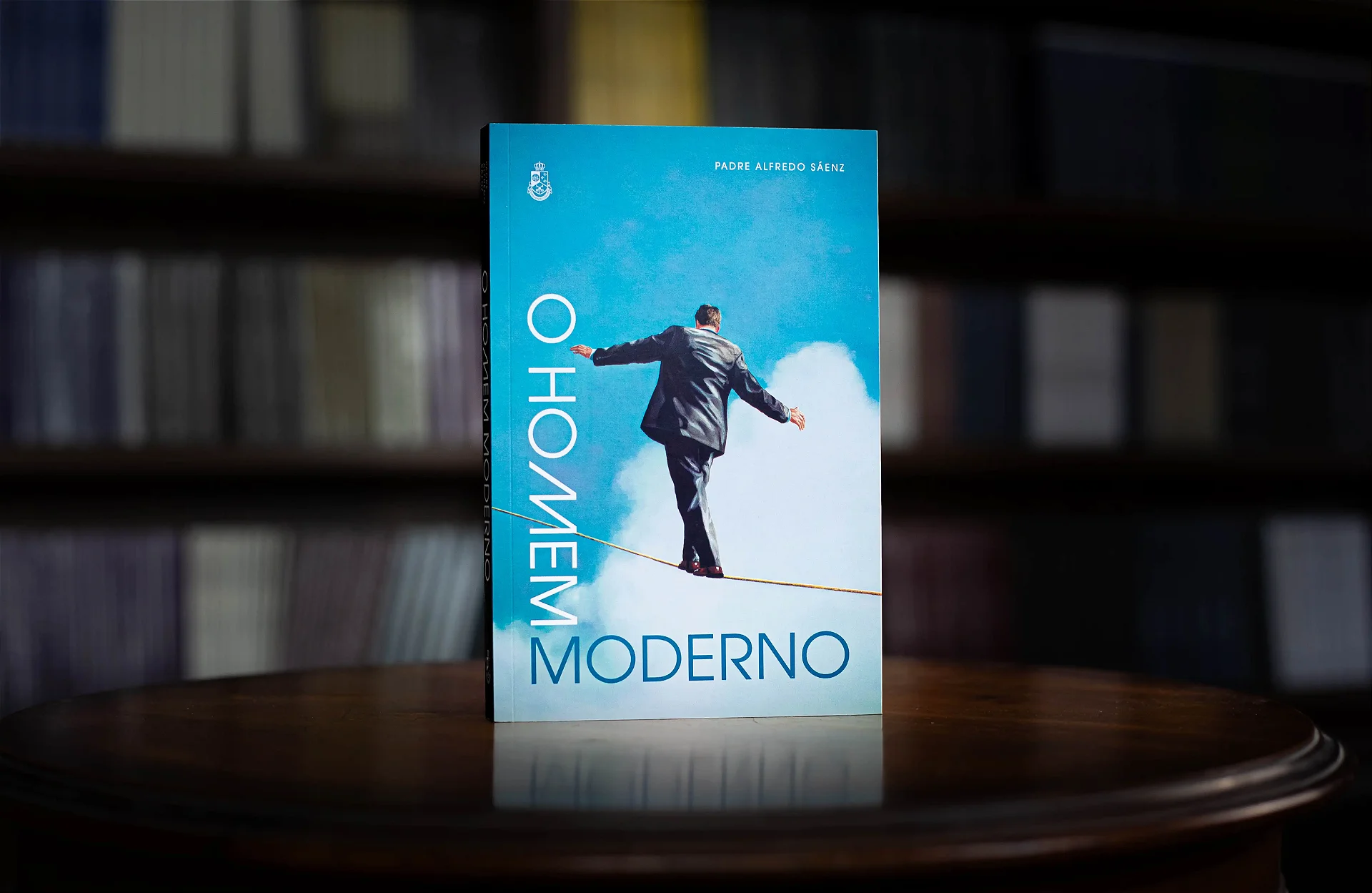 Livro O Homem Moderno em pé sobre a mesa, com estante de livros ao fundo