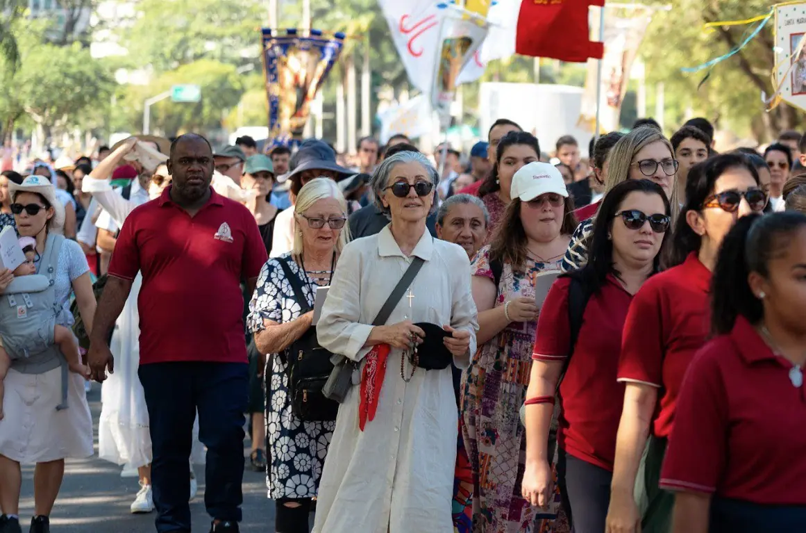 A atriz Cássia Kis e fiéis participam da procissão de Cristo Rei no Rio de Janeiro. Close-up da multidão de fiéis na procissão de Cristo Rei. A atriz Cássia Kis (Mulher de camisa longa bege claro e óculos escuros) caminha entre outros participantes. Ao fundo, bandeiras e estandartes religiosos do cortejo, em um dia ensolarado no Rio de Janeiro.