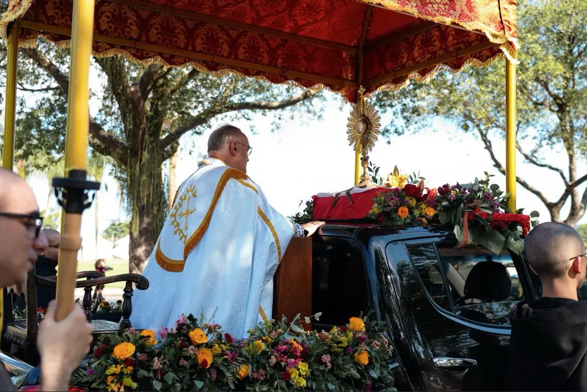 Dom Bernard Fellay em paramentos brancos no carro eucarístico ricamente decorado na procissão de Cristo Rei. Close-up de Dom Bernard Fellay, de costas, vestindo paramentos brancos e dourados, em pé sobre o carro com o Santíssimo Sacramento. O ostensório está visível sob o dossel vermelho e dourado, rodeado por abundantes arranjos florais nas cores amarelo, laranja e vermelho, em uma alameda arborizada.