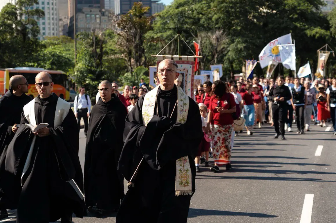 Dom Lourenço Fleichman liderando a procissão de Cristo Rei entre clérigos e fiéis no Rio de Janeiro. Dom Lourenço Fleichman, usando batina preta e estola bordada branca e dourada, lidera um grupo de clérigos vestidos de preto durante a procissão de Cristo Rei. A multidão de fiéis e bandeiras religiosas seguem atrás em uma avenida urbana, com prédios altos e árvores visíveis no fundo.
