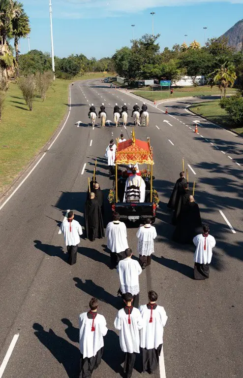 Vista aérea da liderança da procissão de Cristo Rei com cavalaria e clérigos no Rio de Janeiro. Imagem aérea vertical mostrando a vanguarda da procissão de Cristo Rei. O carro com o Santíssimo Sacramento (sob dossel vermelho) é cercado por clérigos de batinas pretas e coroinhas de sobrepeliz branca. A cavalaria da polícia militar segue à frente, abrindo a pista de uma avenida arborizada, com o Pão de Açúcar ao fundo (no canto superior direito).