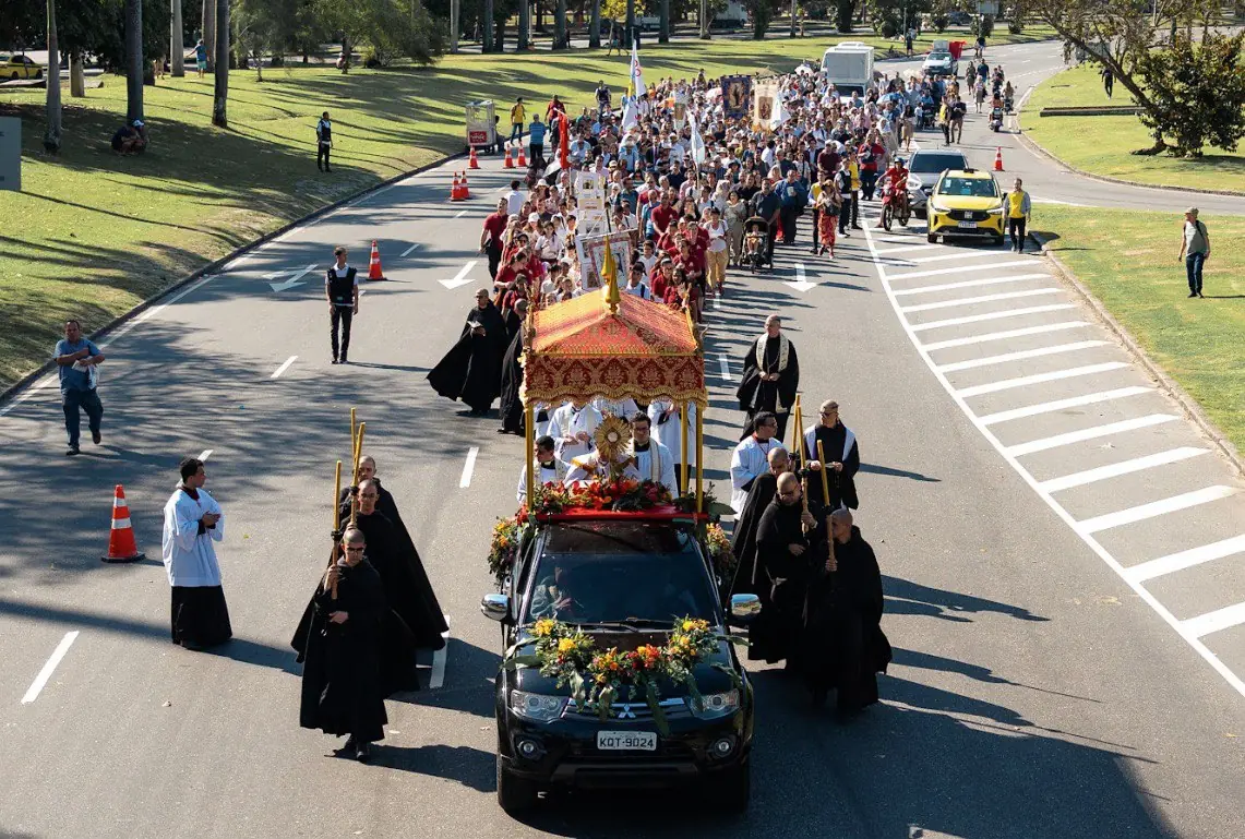 Procissão solene de Cristo Rei em avenida no Rio de Janeiro, com clérigos cercando o Santíssimo Sacramento. Vista de cima da procissão de Cristo Rei em uma avenida larga e arborizada. O veículo com o Santíssimo Sacramento, coberto por um dossel vermelho e dourado e cercado por religiosos de batinas pretas, lidera uma grande multidão de fiéis. O cortejo ocupa a pista da direita da avenida, com trânsito desviado.