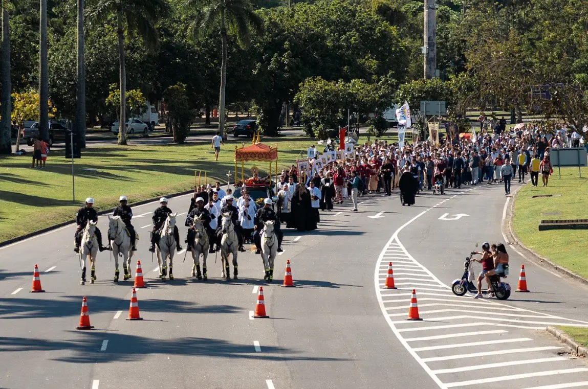Cavalaria abrindo caminho para a procissão de Cristo Rei e grande multidão de fiéis em alameda do Rio de Janeiro. Vista frontal da procissão de Cristo Rei em uma via arborizada (com muitas palmeiras). Seis policiais montados a cavalo lideram, seguidos pelo carro do Santíssimo Sacramento sob um dossel vermelho e uma grande multidão de fiéis a pé. Cones de sinalização e uma motocicleta com passageiros na lateral da pista indicam o bloqueio da via para o evento.