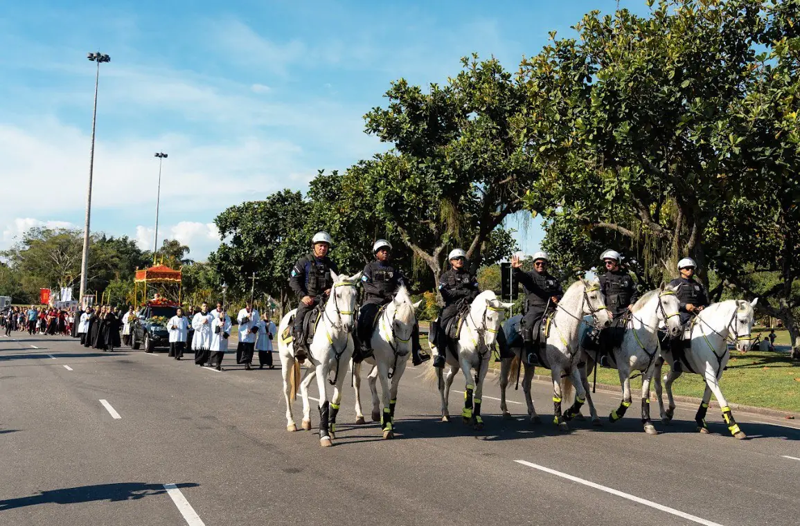 Cavalaria da Polícia Militar e procissão de Cristo Rei em longa vista no Rio de Janeiro. Uma procissão religiosa vista de longe, com seis policiais militares montados em cavalos brancos à direita, liderando o cortejo. Ao fundo, o carro com o Santíssimo Sacramento sob um dossel, seguido por clérigos e fiéis. Uma alameda arborizada se estende ao longo da via urbana sob um céu azul.