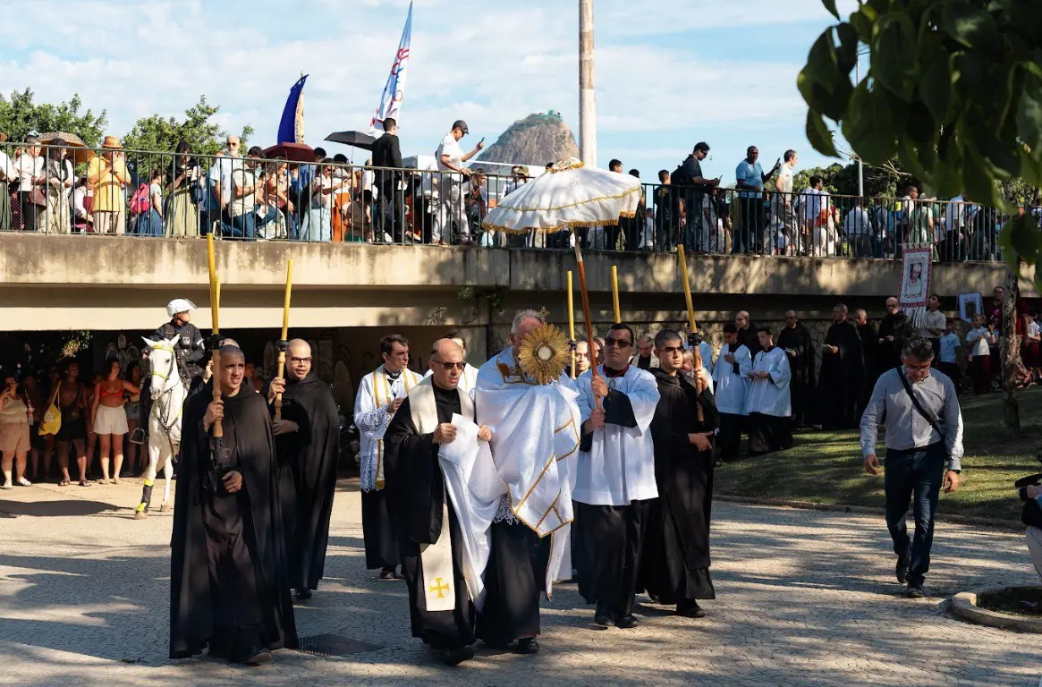 Clérigos e Dom Fellay conduzindo o Ostensório a pé, sob pálio, durante procissão de Cristo Rei. Parte da procissão de Cristo Rei com clérigos a pé. Um padre (possivelmente Dom Fellay) em paramentos brancos, sustentando o Ostensório com o Santíssimo Sacramento sob um pálio (guarda-chuva cerimonial) branco. É cercado por clérigos de batinas pretas. Fiéis observam a passagem da procissão de cima de uma passarela/ponte.