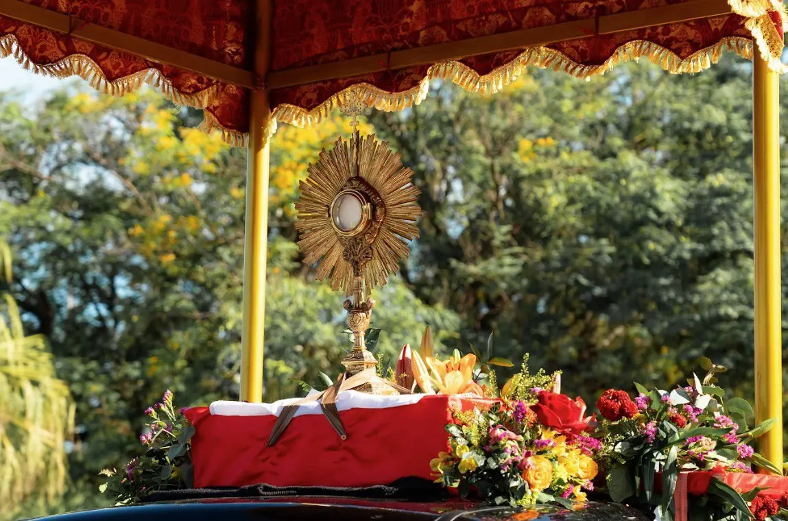Close-up do Santíssimo Sacramento no Ostensório, durante procissão solene de Cristo Rei. Close-up do Ostensório dourado com o Santíssimo Sacramento (hóstia consagrada), colocado sobre um suporte vermelho decorado com ricos arranjos de flores vermelhas, amarelas e laranjas. O ostensório está sob o dossel (baldaquino) vermelho e dourado do carro, com folhagens verdes e céu azul ao fundo.