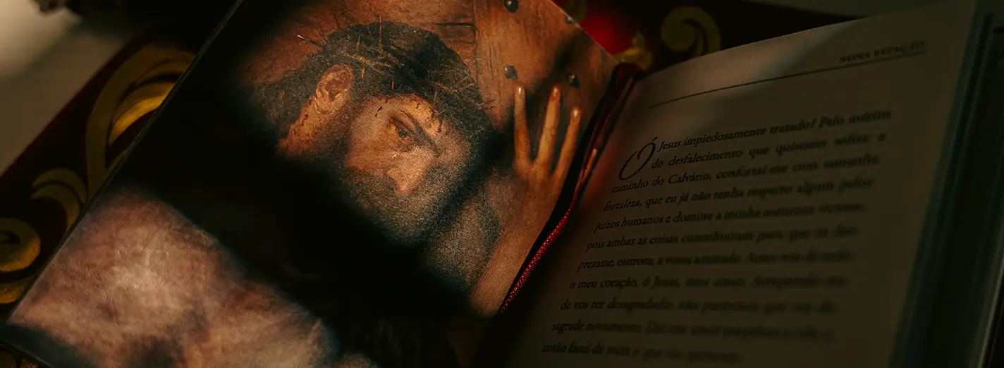 Livro A Via Sacra Ilustrada aberto em uma imagem que mostra o olhar de Jesus coroado de espinhos e carregando a cruz