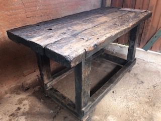 Antiga mesa de fazenda, rústica, feita em tora de madeira com detalhes em ferro na lateral, uma prateleira, mesa de apoio